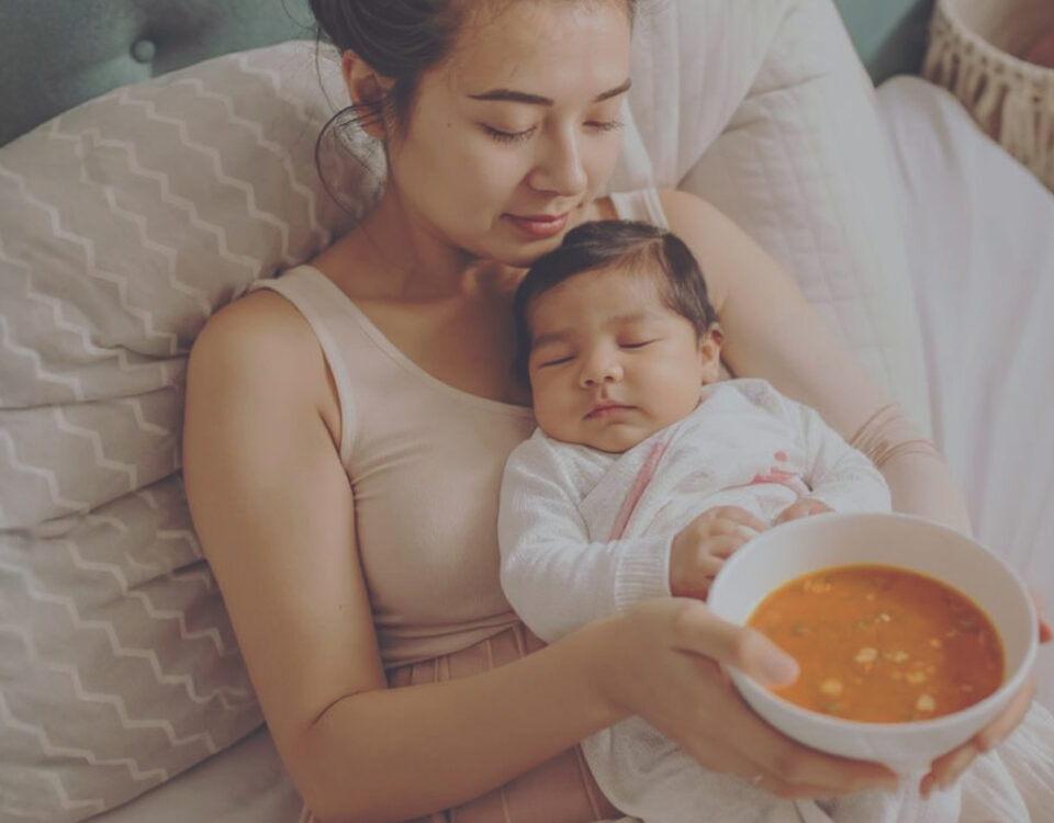 Frisch gebackene Mutter kuschelt mit ihrem Baby, während eine gesunde Mahlzeit von Mothers Finest bereitsteht.